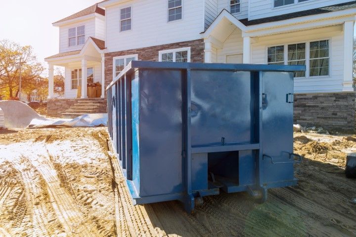 A large blue dumpster sits on the dirt ground in front of a house under construction.