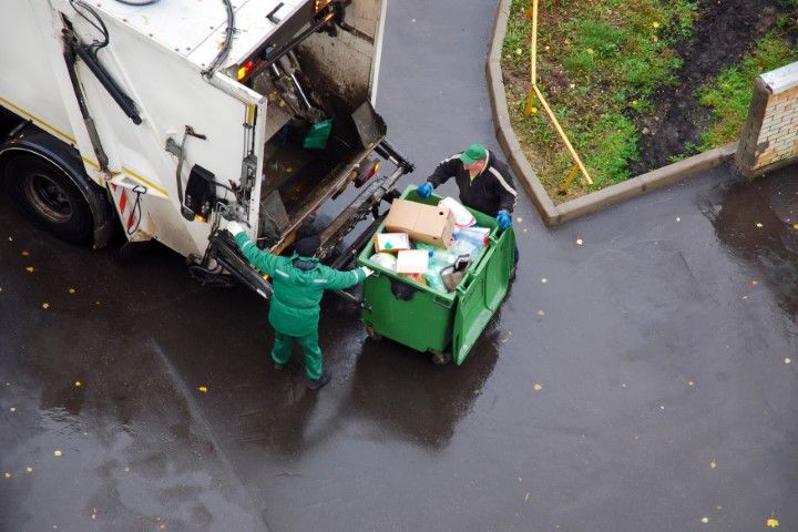 Two workers in uniforms empty a green dumpster into a white garbage truck on a rainy paved surface.