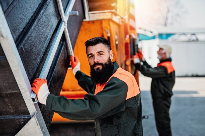 Two sanitation workers in high-visibility uniforms operate a waste collection vehicle outdoors.