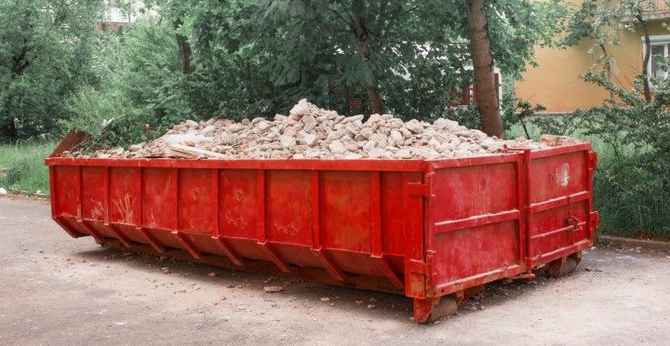 A large, red, rectangular industrial dumpster filled with debris and rubble, parked on a paved lot near some trees.
