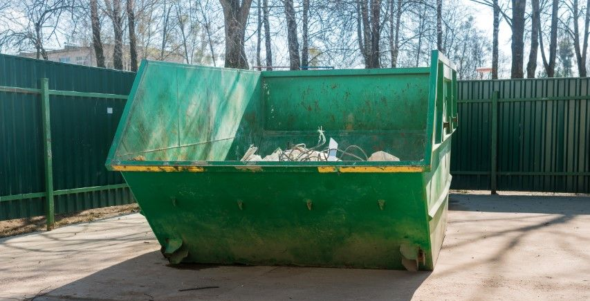 A large, empty green metal dumpster sitting on a paved surface in front of a green privacy fence and bare trees.