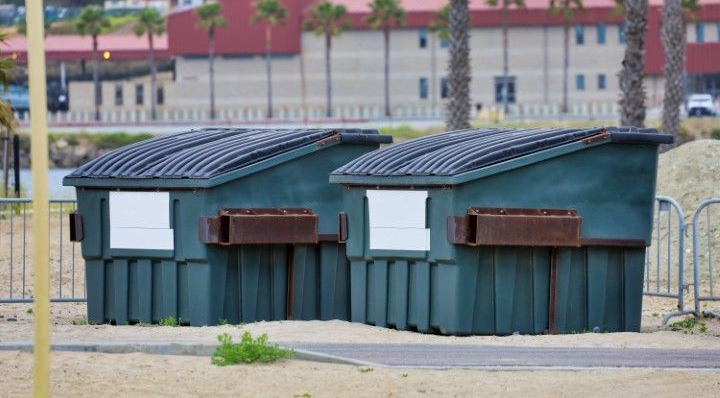 Two dark green dumpsters with black lids sit side-by-side on a sandy ground in front of a metal fence and palm trees.