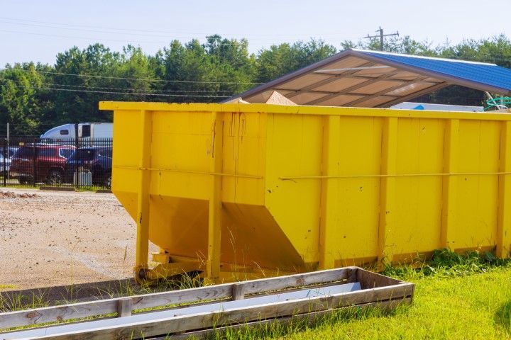 A large, yellow industrial dumpster sits on a gravel lot next to a low wooden structure under a blue-roofed shelter.