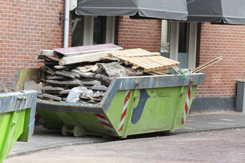 A lime-green construction skip filled with debris and wooden pallets parked on a street in front of a brick building.