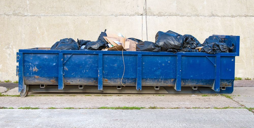 A long blue dumpster filled with black trash bags and cardboard boxes sits against a light concrete wall.