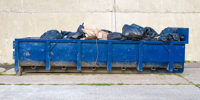 A long blue dumpster filled with black trash bags and cardboard boxes sits against a light concrete wall.
