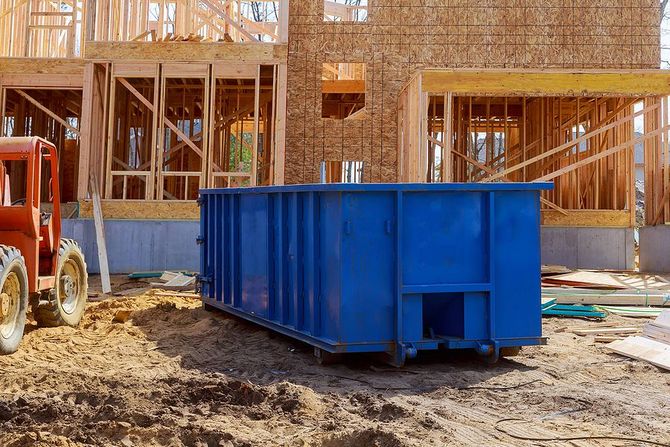 A blue construction dumpster parked on a dirt lot in front of a house under construction with visible wooden framing.