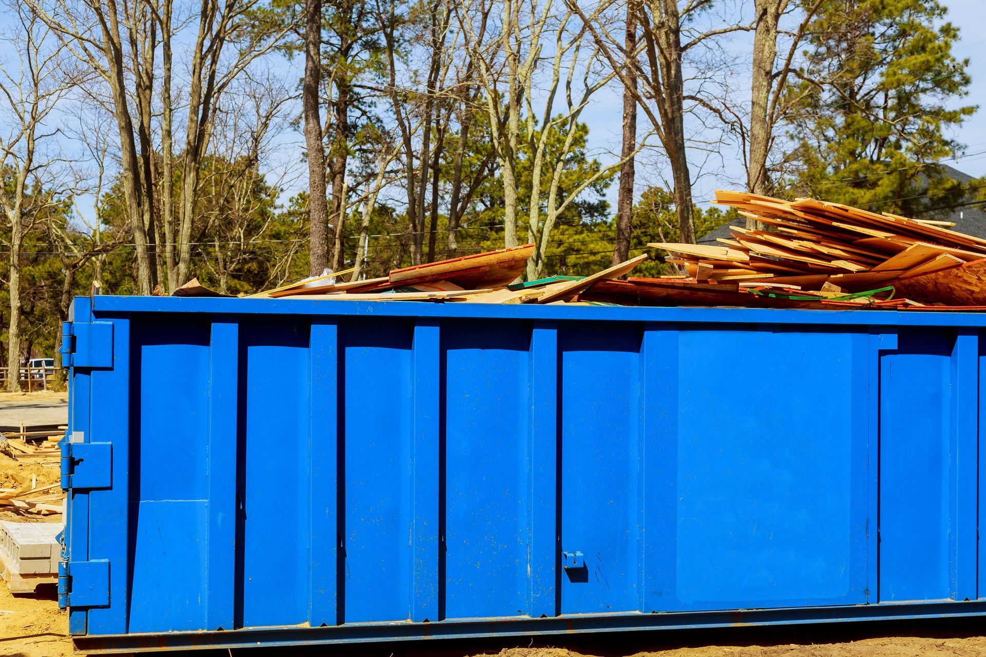 A large, bright blue industrial dumpster filled with scrap wood sits outdoors in a wooded area.