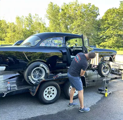 Man working on a black race car on a trailer in a sunny outdoor lot