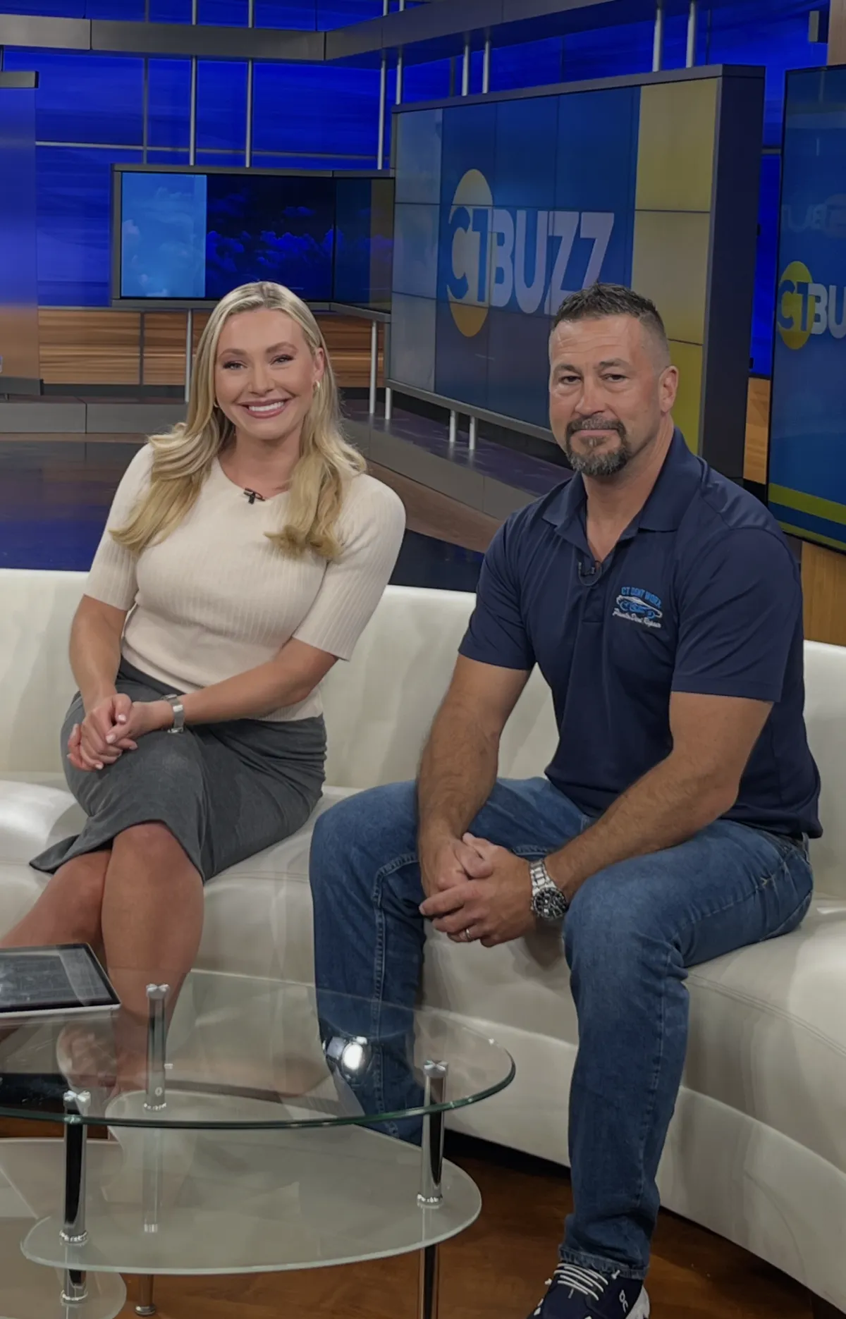 Two people seated in a TV studio with a blue “BUZZ” backdrop, smiling at the camera.