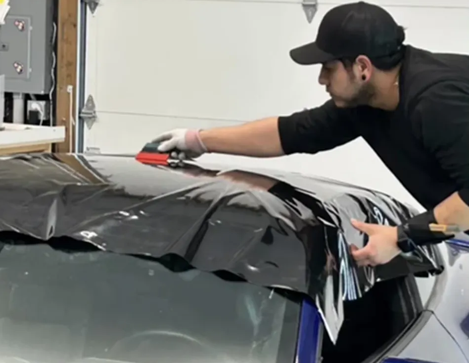 Technician applying black vinyl wrap to a car roof in a workshop