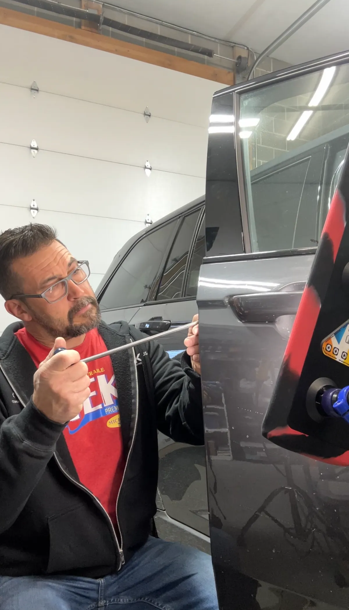 Man in glasses works on a black car in a garage, using a tool near the side panel.