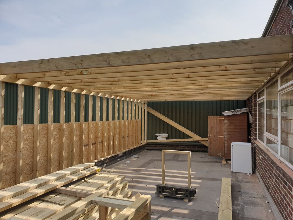 Construction of a wooden carport with OSB walls and corrugated metal backing, next to a brick building.