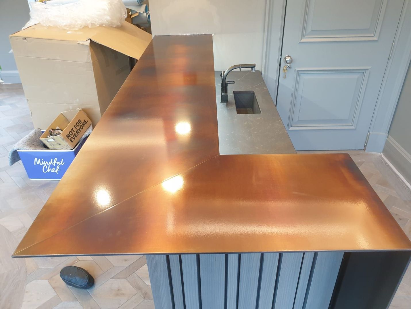 Copper-colored L-shaped countertop on a grey and white cabinet, with a sink and faucet in the background.