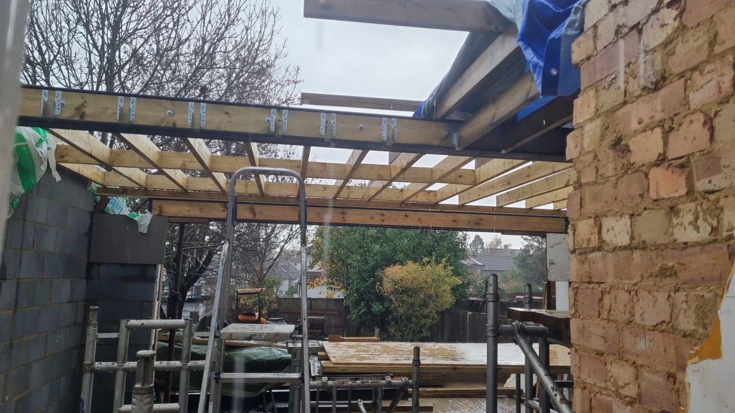 Construction site with exposed wooden beams, partially built roof. Brick and cinder block walls, ladder visible.