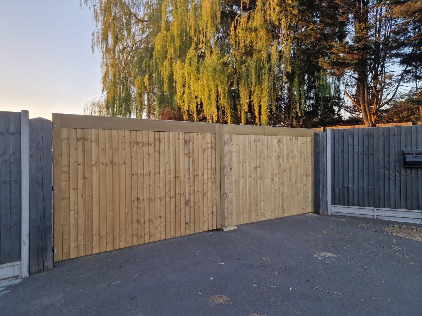 Wooden driveway gates, light brown, set between gray concrete posts on paved surface, with trees in background.