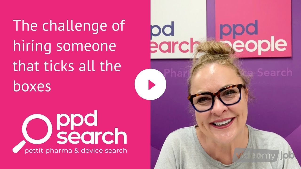 A woman in glasses smiles in front of a pink PPD Search sign, discussing hiring.