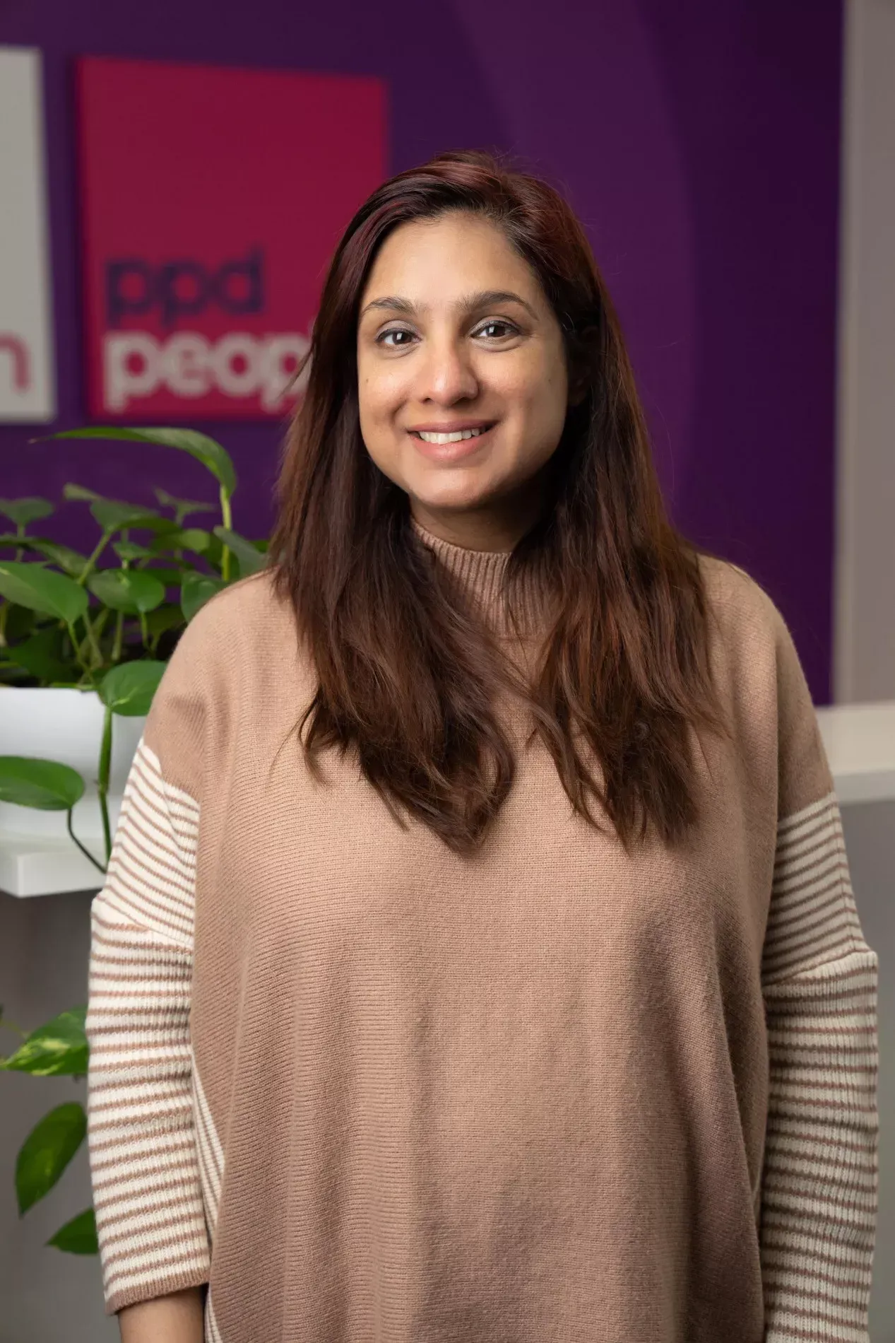 Woman with long brown hair smiles. She wears a tan sweater with striped sleeves. Purple wall with logo behind her.