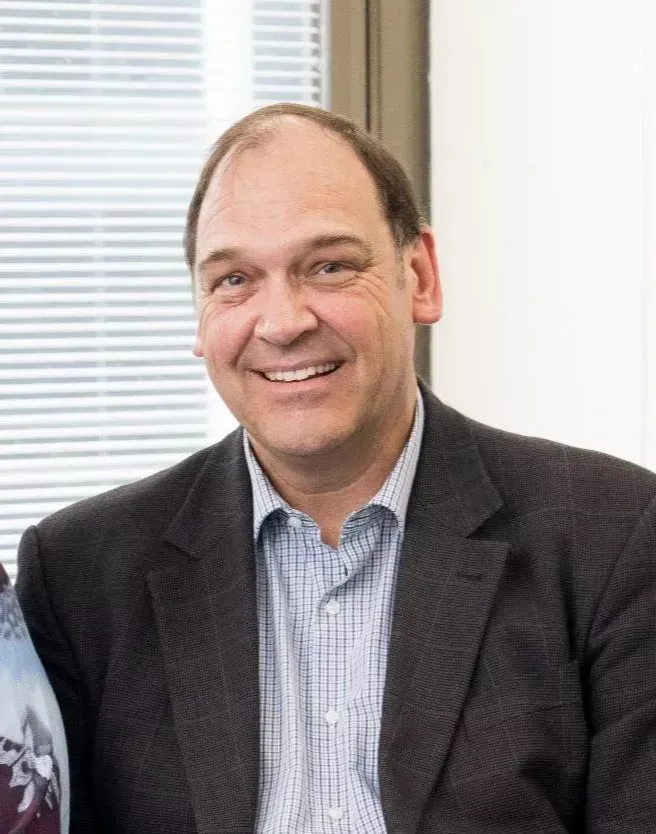 Man with a warm smile, wearing a dark suit jacket over a light blue collared shirt, in an office.