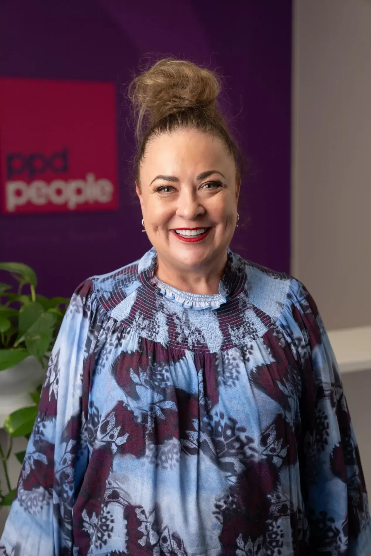 Woman with a bun smiling; in front of a purple “pod people” sign. Blue and purple patterned blouse.