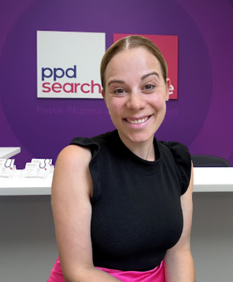 Woman smiling at camera in black top and pink skirt, purple wall behind, with PPD Search logo visible