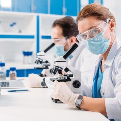 Two lab workers using microscopes, wearing masks, goggles, and gloves. White coats, blue lab setting.