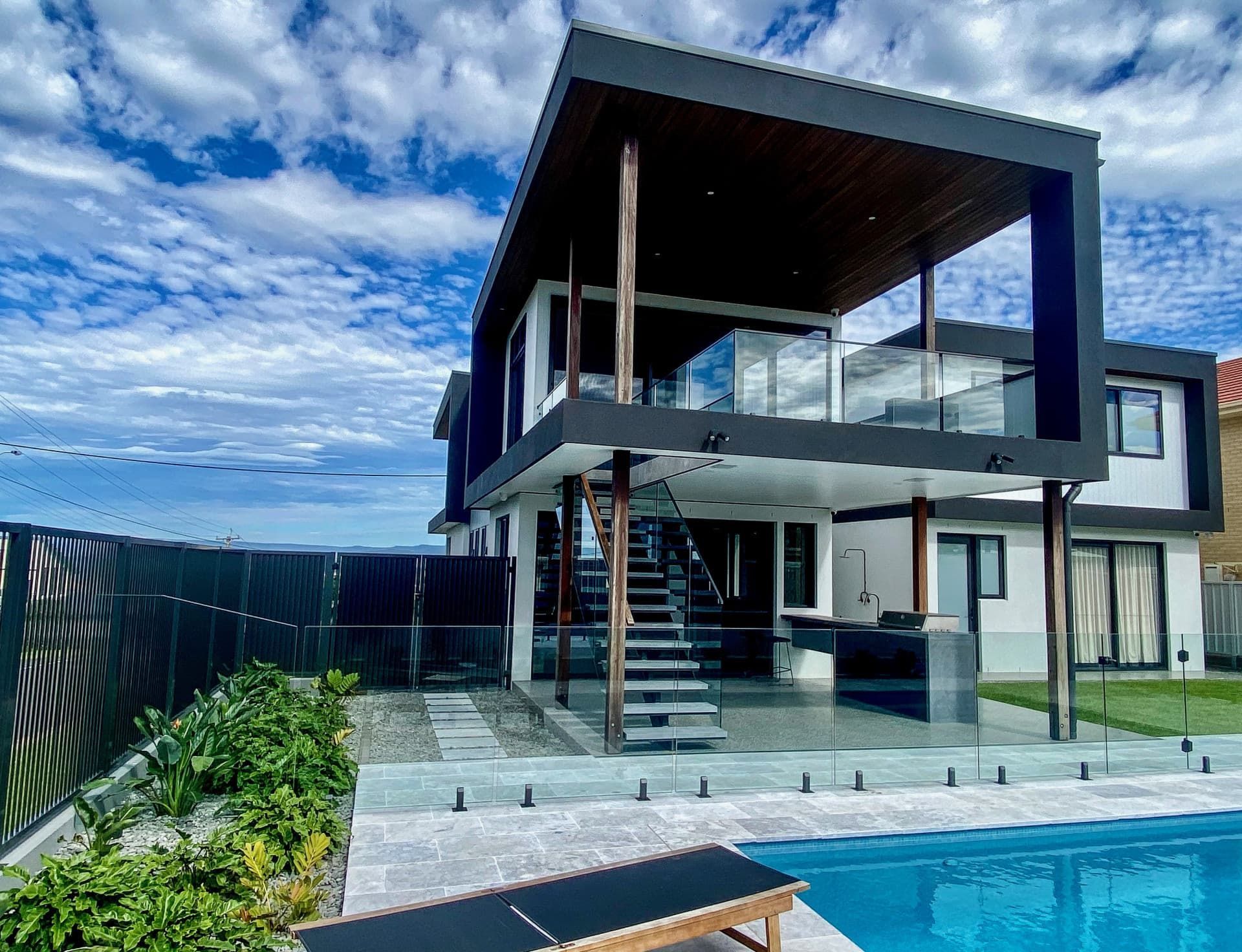 Modern two-story house with a black frame over an outdoor area, pool in the foreground, cloudy sky — Envisage Construction and Design in Mountain, NSW