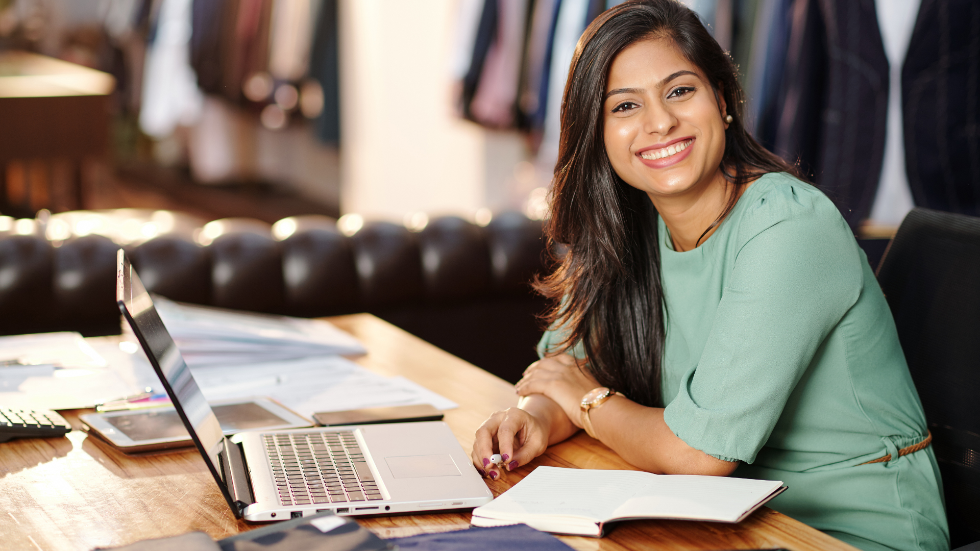 A smiling person sits at a desk with a laptop and documents in a professional clothing boutique.