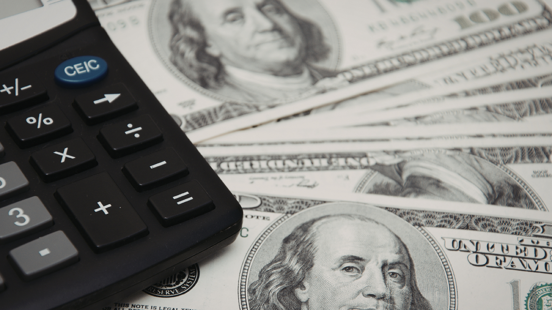 A black calculator sits atop a small fan of United States one-hundred-dollar bills.