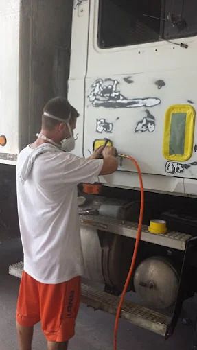 Man sanding the side of a white truck. He wears a mask, orange shorts, and is in a shop.