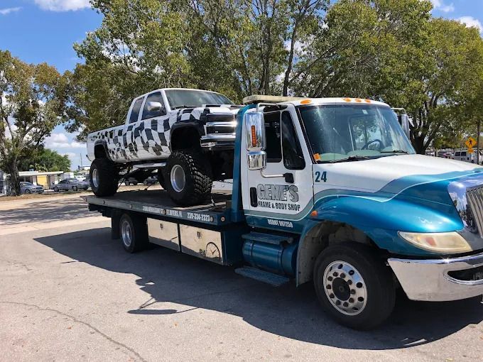 A lifted truck with camouflage paint is towed on a flatbed tow truck on a sunny day.