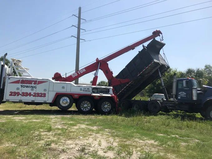 Tow truck lifting a blue dump truck's bed in a grassy area with power lines in the background.