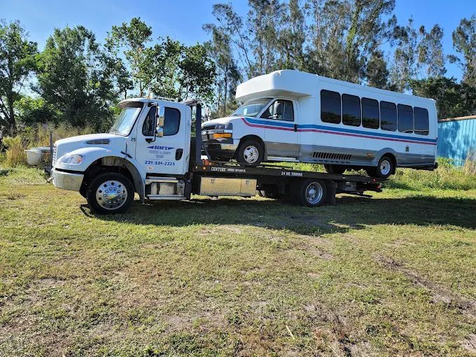 Tow truck carrying a white and blue bus on a grassy field under a blue sky.