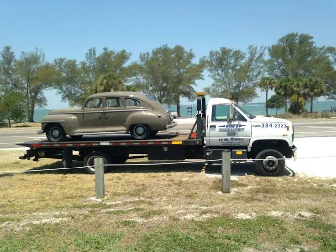 Tan classic car on a flatbed tow truck. Seaside setting with blue sky and trees.