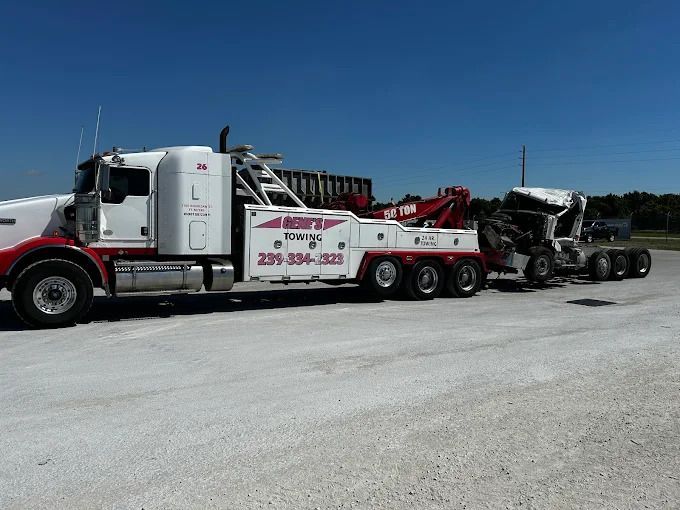 A white tow truck with red accents tows a damaged semi-truck on a paved lot under a blue sky.
