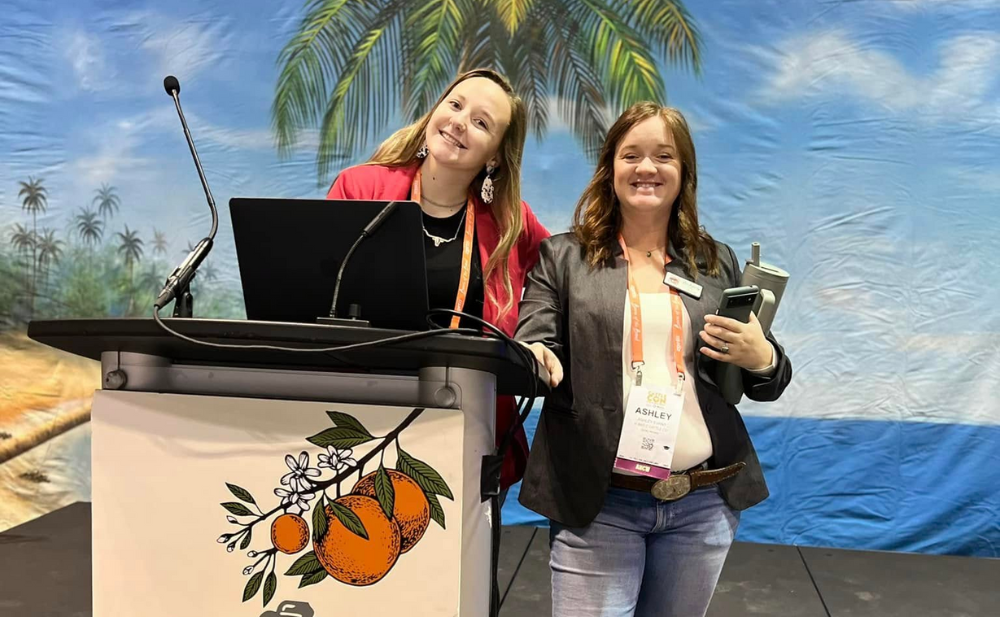 Two women smiling by podium with laptop and microphone, orange logo. Tropical background.