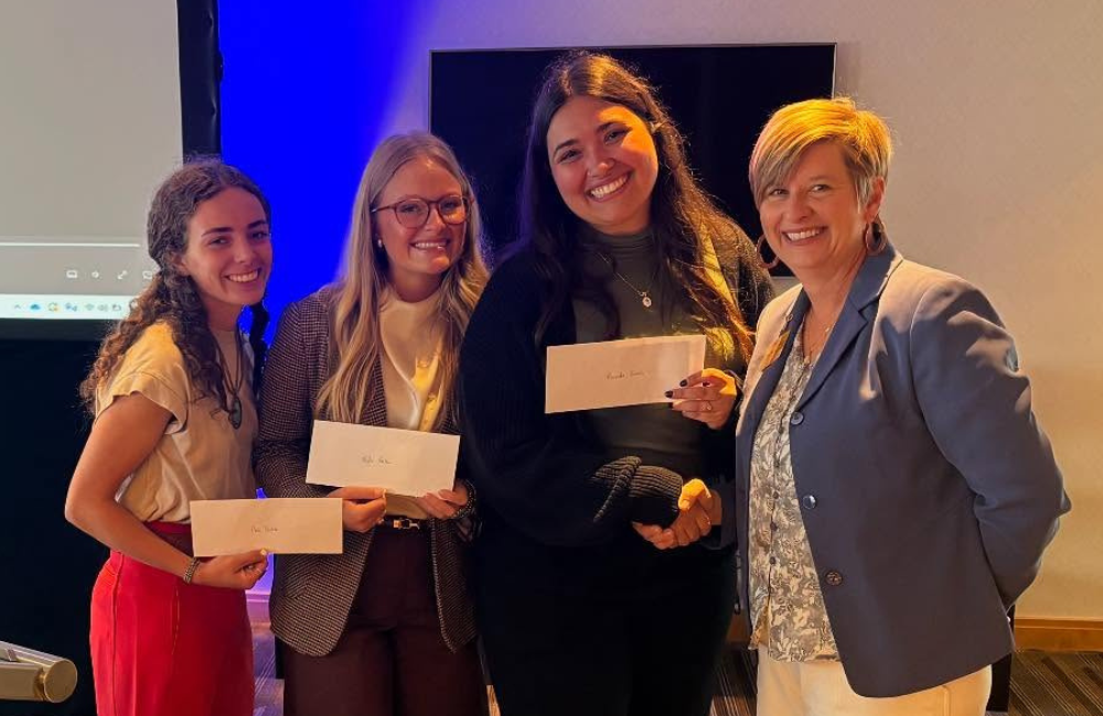 Four women holding checks, smiling at camera. Award presentation, inside with a blue background.