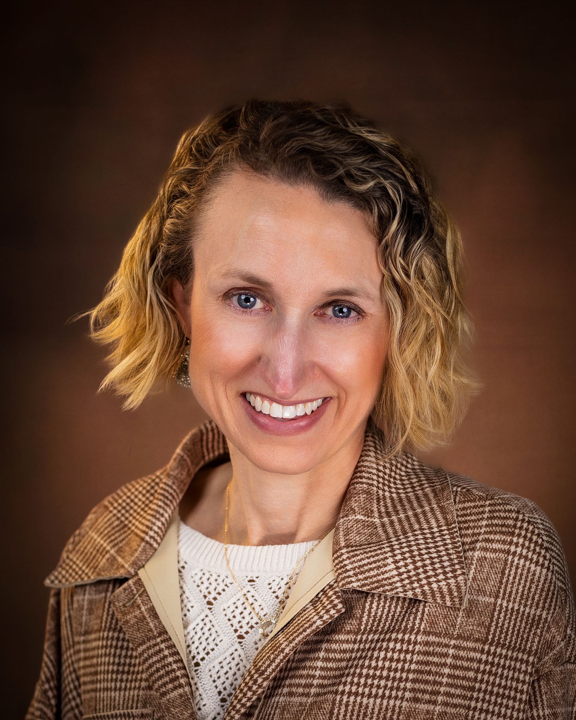 Woman with curly blonde hair smiles, wearing a brown patterned jacket over a white textured sweater. Brown background.