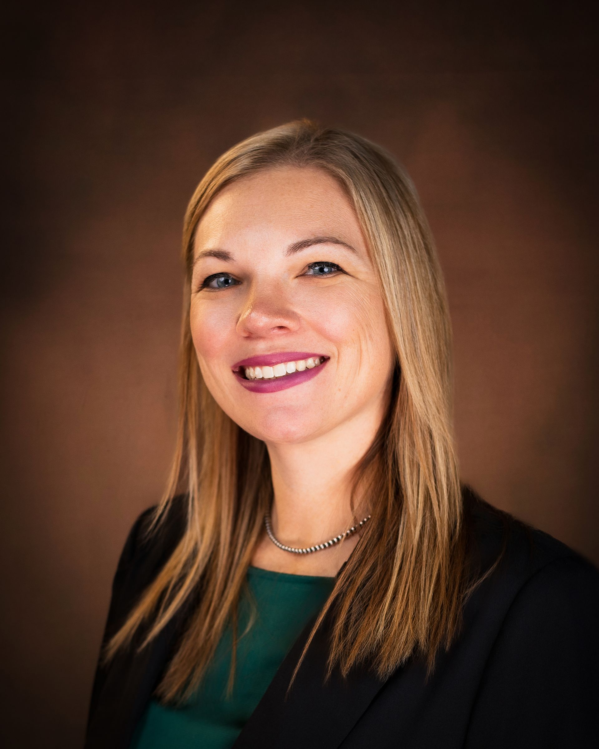 Woman with blonde hair smiling, wearing a dark blazer and green shirt, brown background.