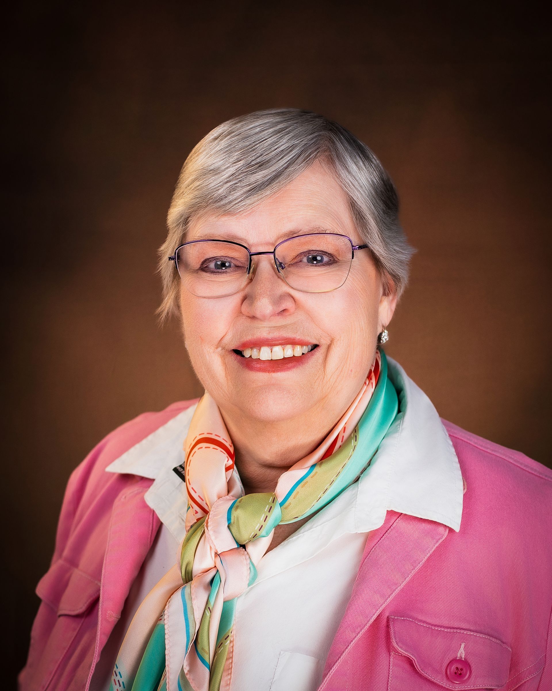 Smiling woman with short gray hair, glasses, and a pink jacket against a brown background.