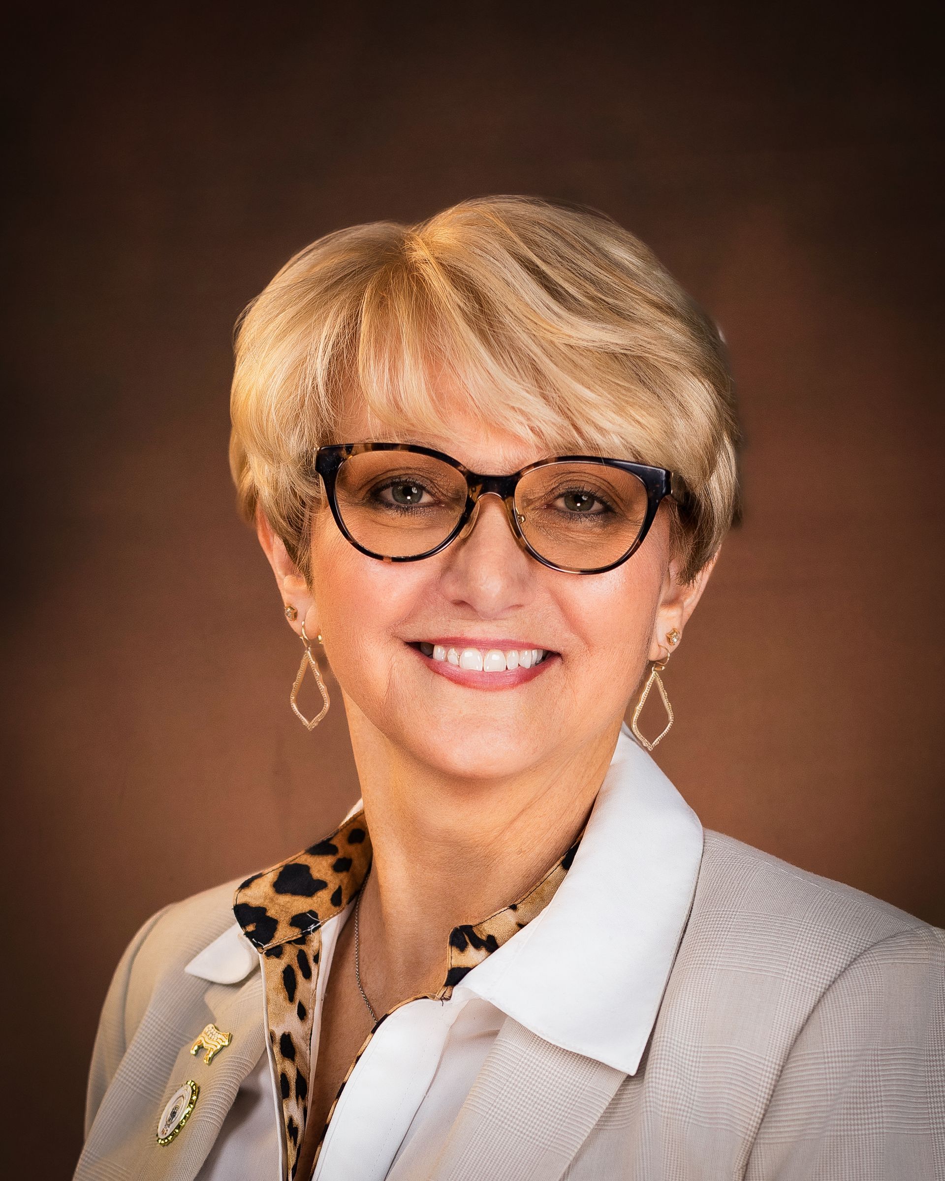 Woman with blonde hair, glasses, smiling, wearing a beige blazer and leopard print shirt.