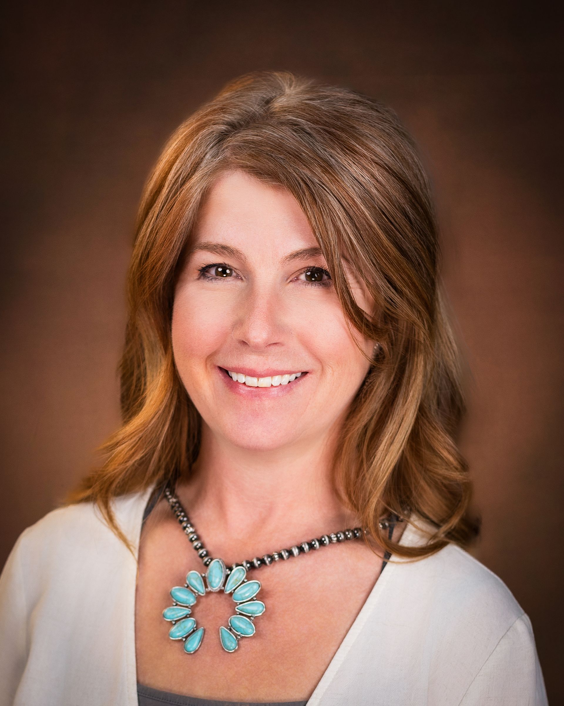 Woman with long brown hair wearing a turquoise pendant necklace, smiling, against a brown background.