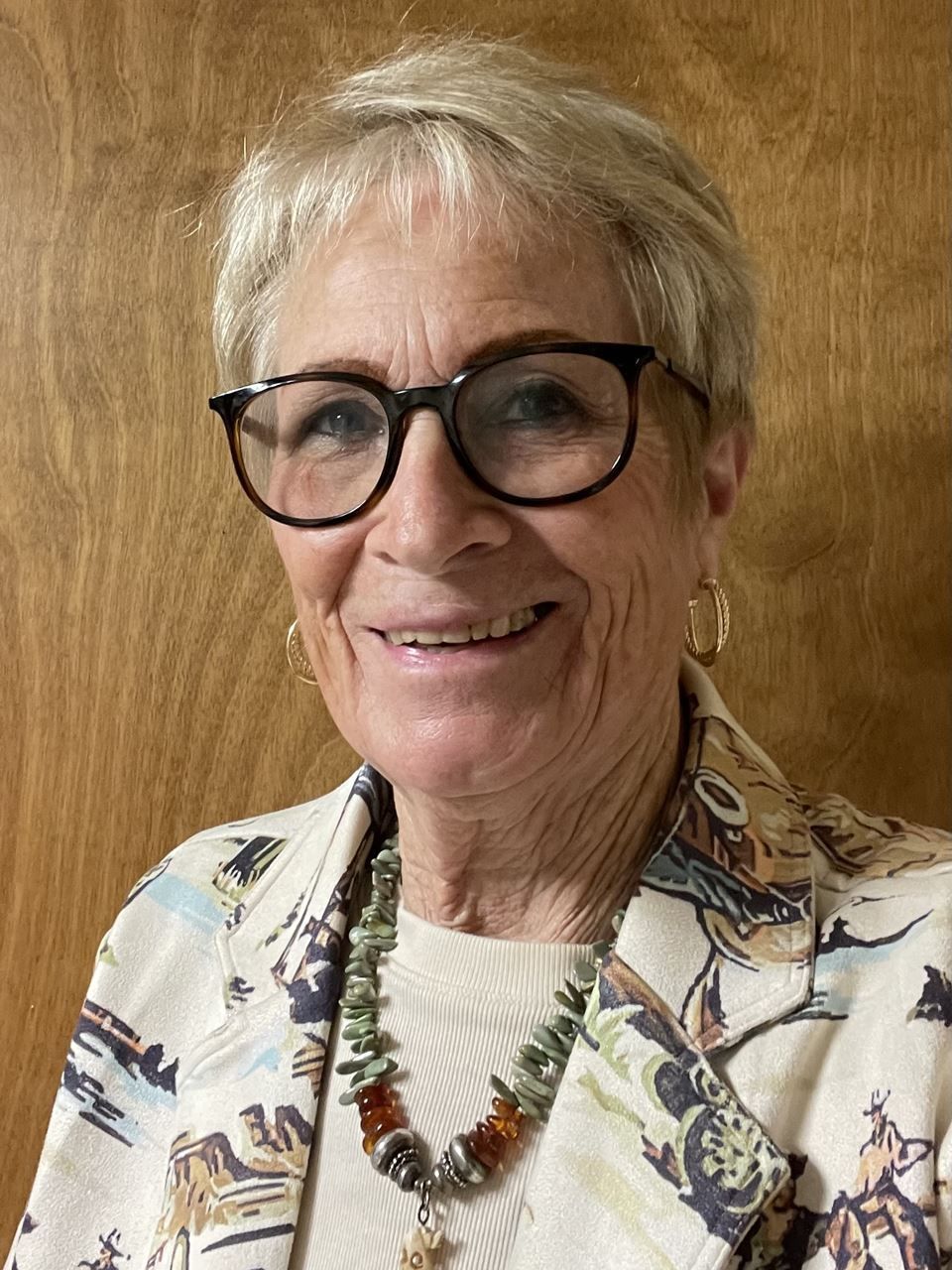 Woman with short, blonde hair, glasses, and a patterned jacket smiles, looking at the camera.