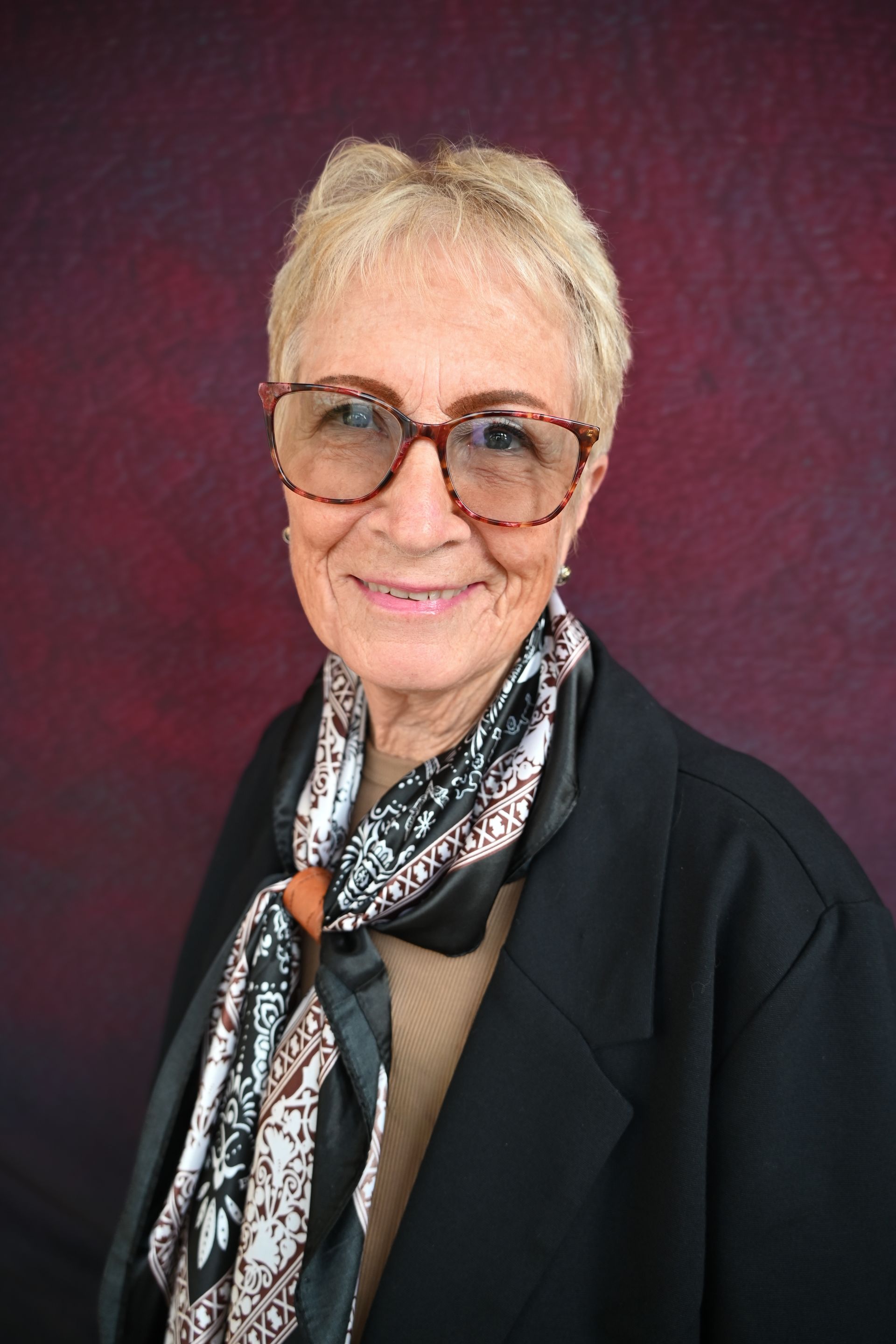 Woman with short, blonde hair, glasses, and a patterned jacket smiles, looking at the camera.