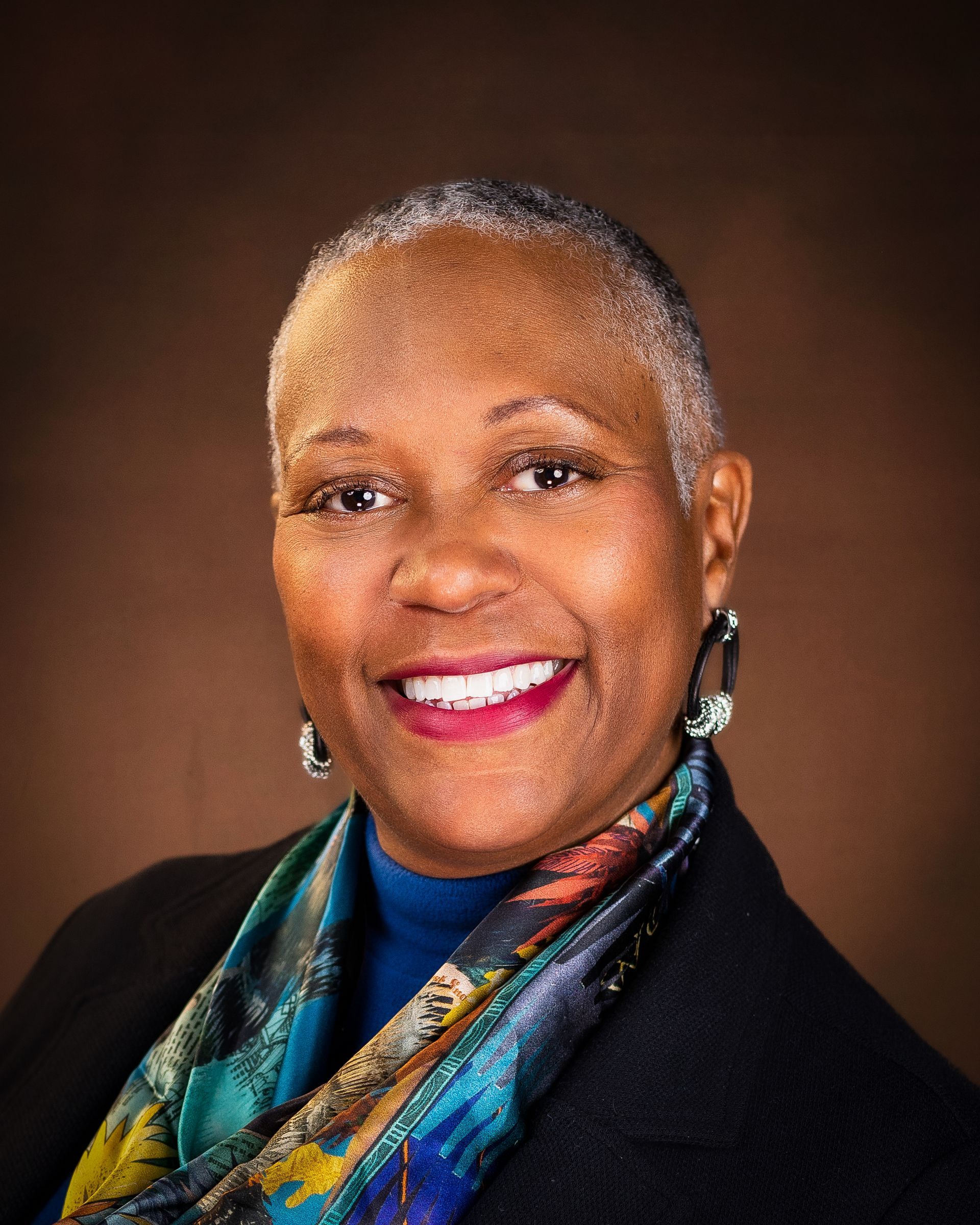 Smiling Black woman with short gray hair, wearing a blue top, scarf, and earrings, in front of a brown backdrop.