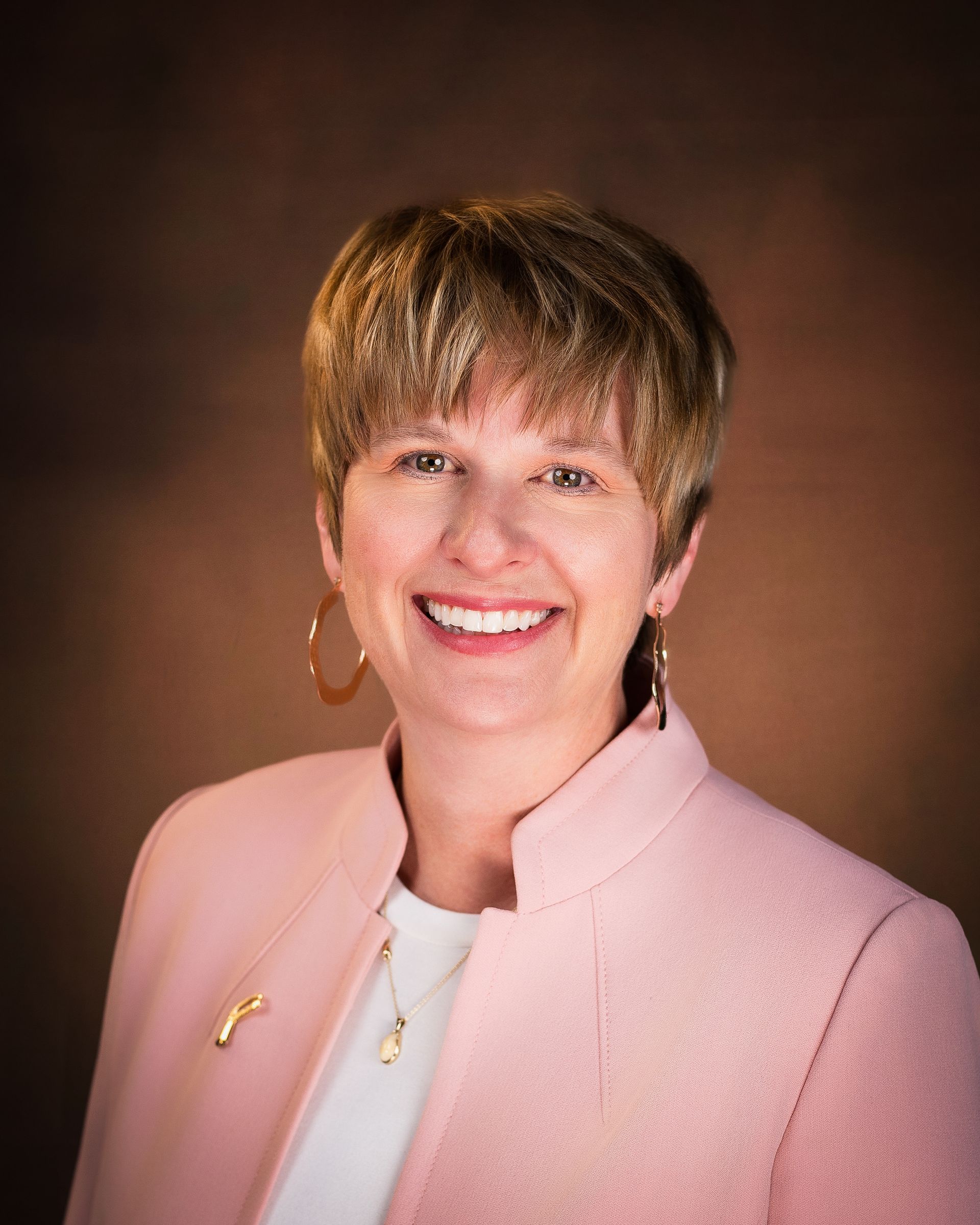 Woman with short blonde hair smiles, wearing a pink jacket and gold hoop earrings; brown background.
