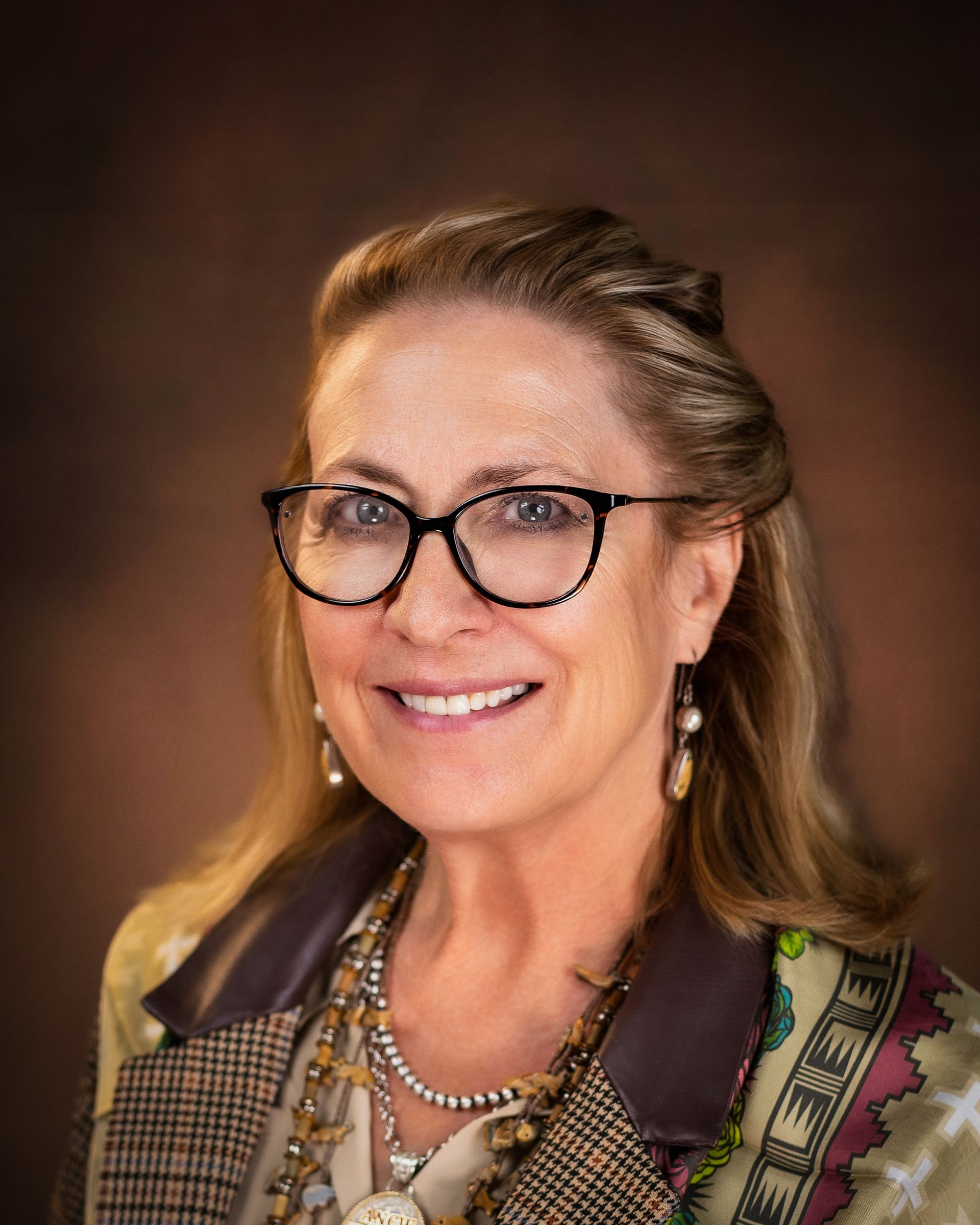 Woman with glasses smiling, wearing a jacket and jewelry, brown background.