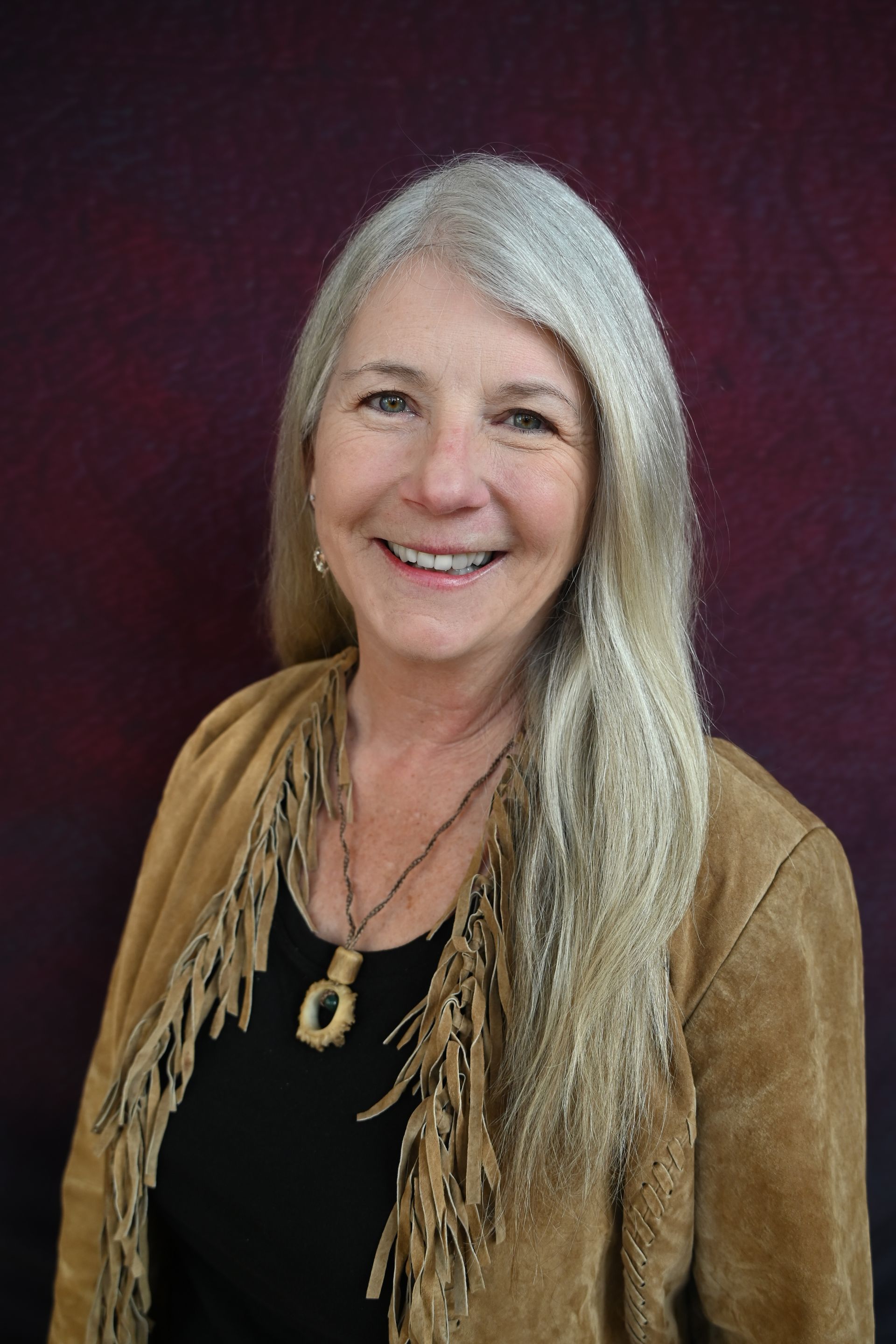 Woman with long brown hair wearing a turquoise pendant necklace, smiling, against a brown background.