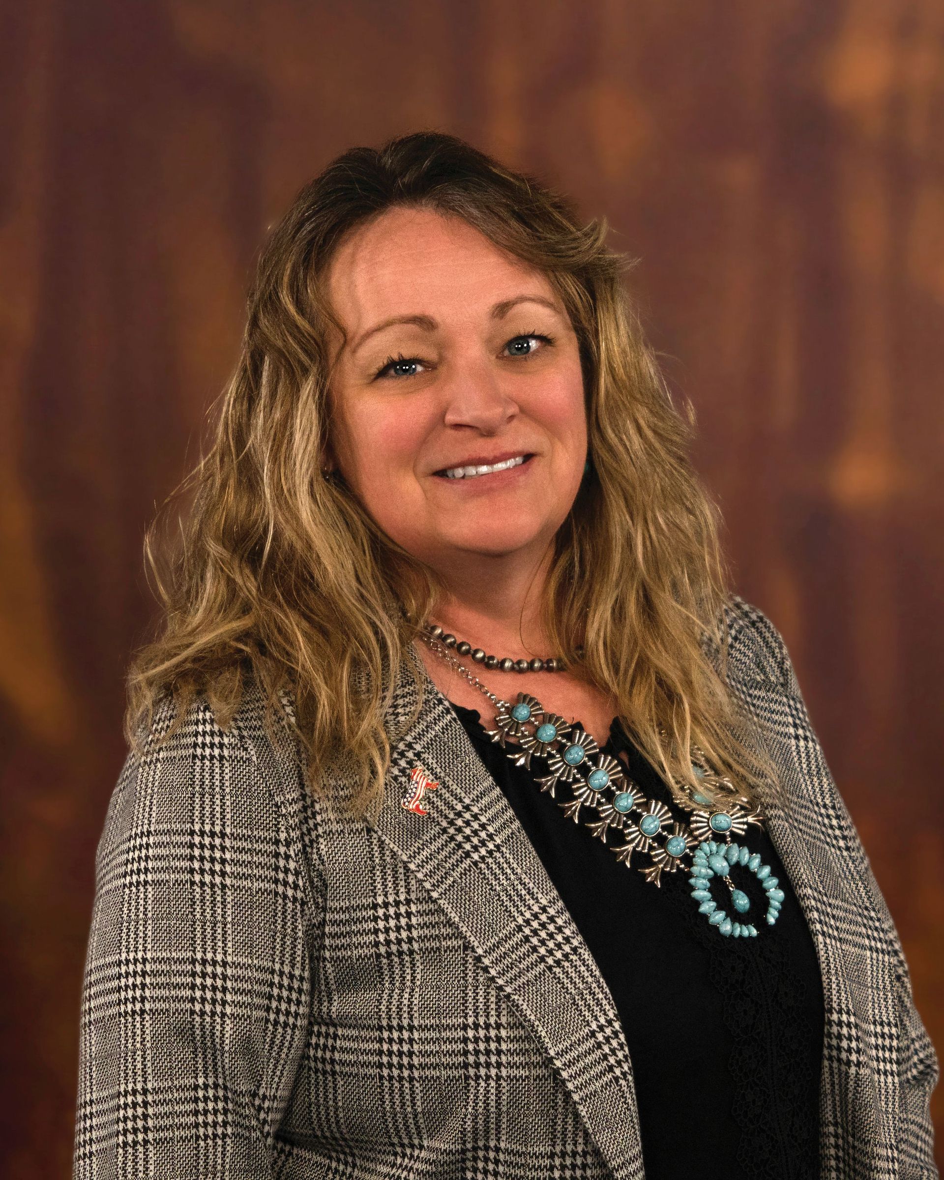 Woman in a blazer and turquoise necklace smiles, against a brown background.
