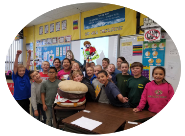 Students in a classroom posing with a giant burger prop, smiling, and cheering near a whiteboard.